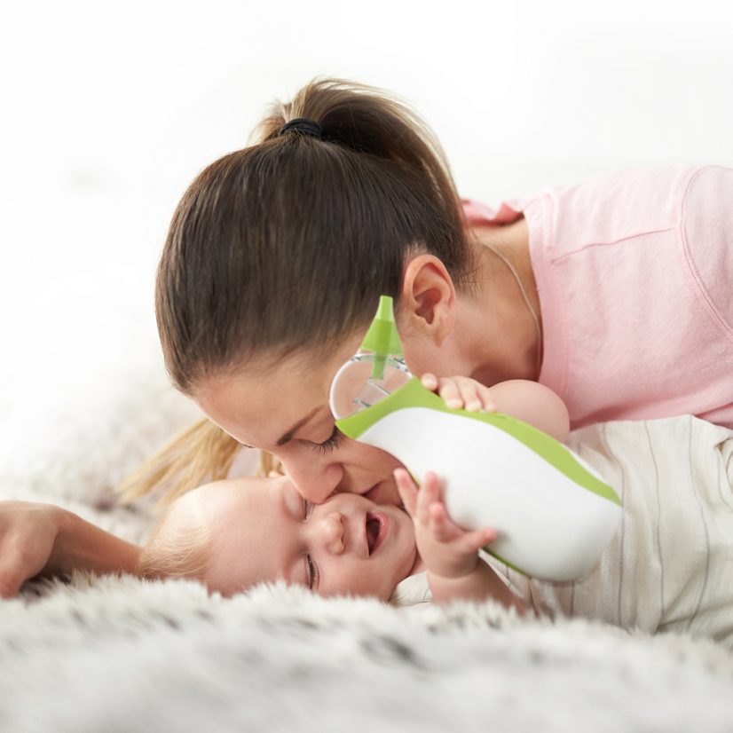 A mom kissing her baby boy who is carrying the Nosiboo Go portable nasal aspirator in his hands while lying on the bed