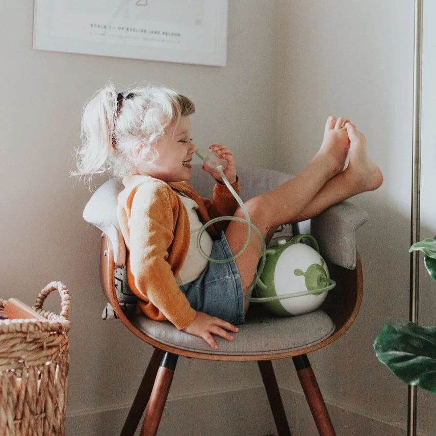 A smiling little girl sitting in a chair holding a Nosiboo Pro electric nasal aspirator in her hands