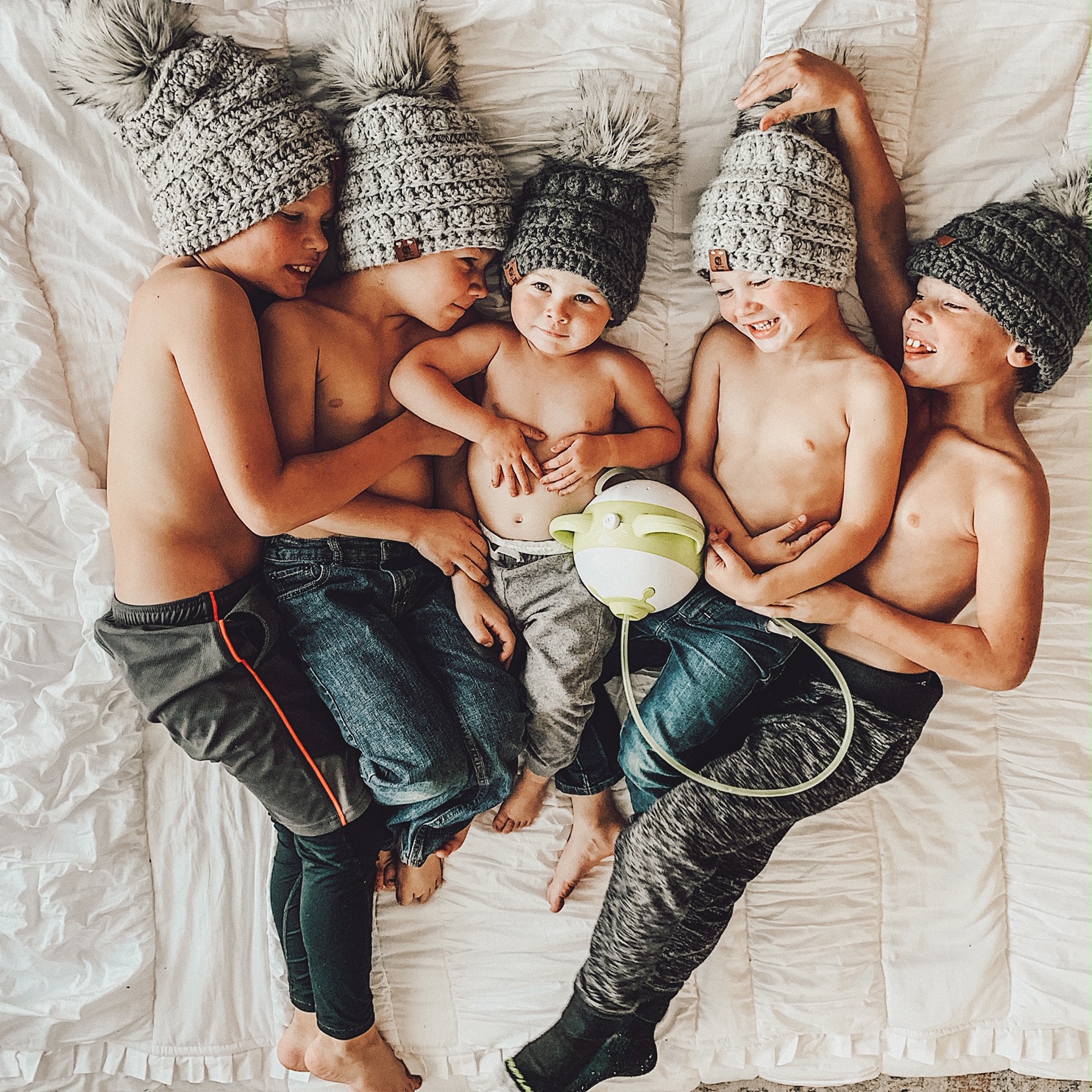 5 smiling brothers lying on a bed next to each other in matching winter hats, with a Nosiboo Pro electric nasal aspirator between them