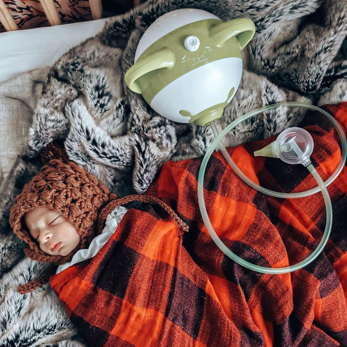 A newborn sleeping in a bed, next to a Nosiboo Pro electric nasal aspirator